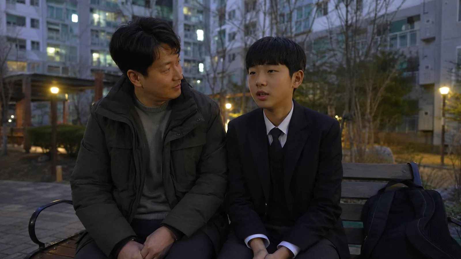 저녁 무렵 아파트 단지 벤치에 나란히 앉아 이야기하는 한국인 아버지와 교복 입은 아들