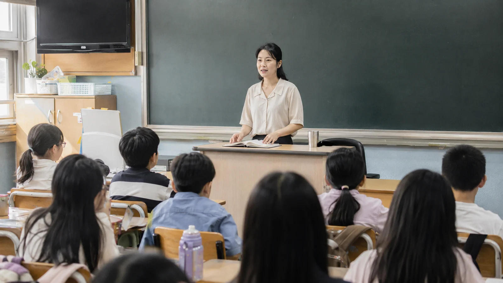 교탁 앞에서 학급 학생들에게 이야기하는 한국인 여자 담임교사와 학생들의 뒷모습