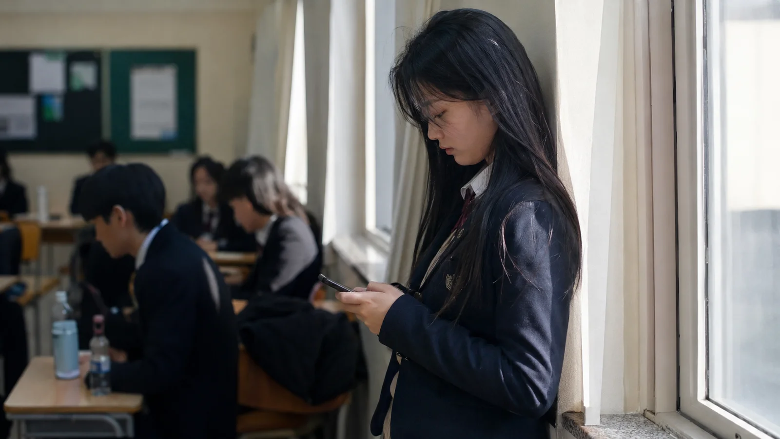쉬는 시간 교실 뒤편 창가에서 혼자 휴대전화를 보는 여고생